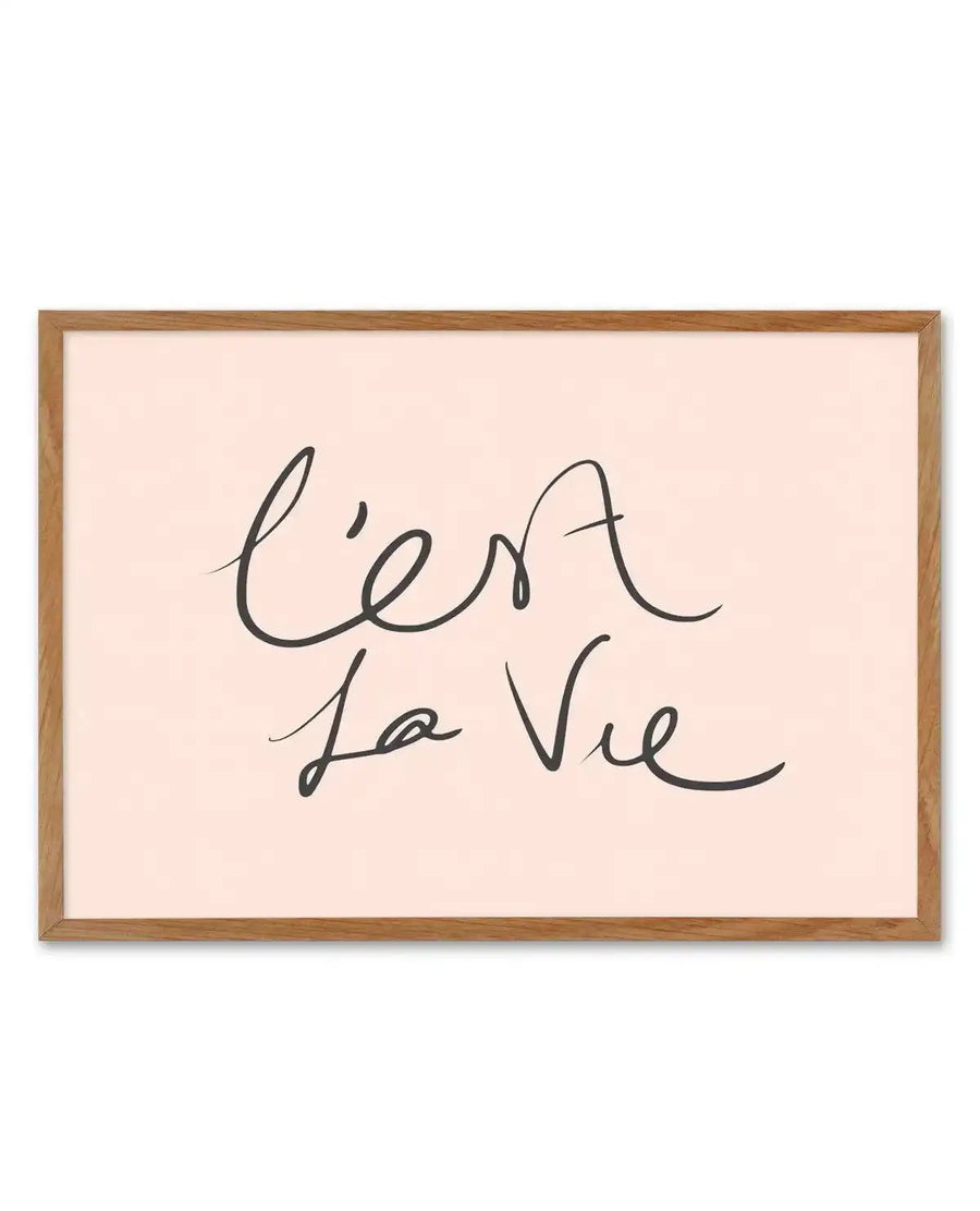 Framed art print featuring handwritten script text reading "C'est La Vie" in dark gray ink on a soft blush pink background. Natural wood frame with white border. Horizontal rectangular composition with flowing, cursive lettering in a casual, elegant style.