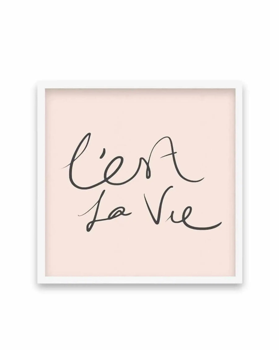 Square art print with white frame displaying handwritten script reading "C'est La Vie" in black ink on a soft, warm beige background with subtle peachy undertones. The lettering is loose and flowing in a casual cursive style. The print has a minimalist aesthetic with generous white space around the text.