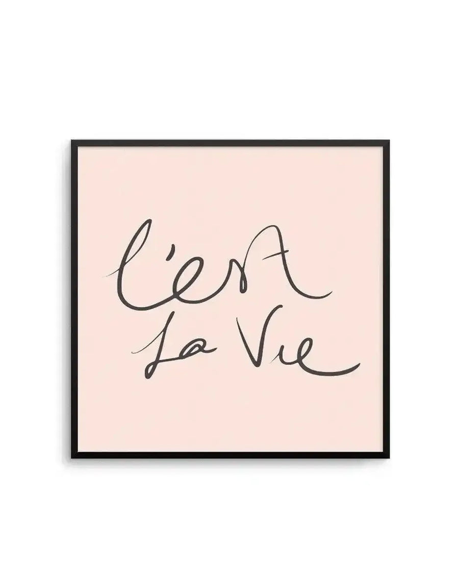 Square art print with a black frame displaying handwritten script reading "C'est La Vie" in dark gray cursive lettering on a soft blush pink background. The casual, flowing handwriting style gives the piece a personal, intimate quality with organic pen strokes and slight variations in line weight typical of natural handwriting.