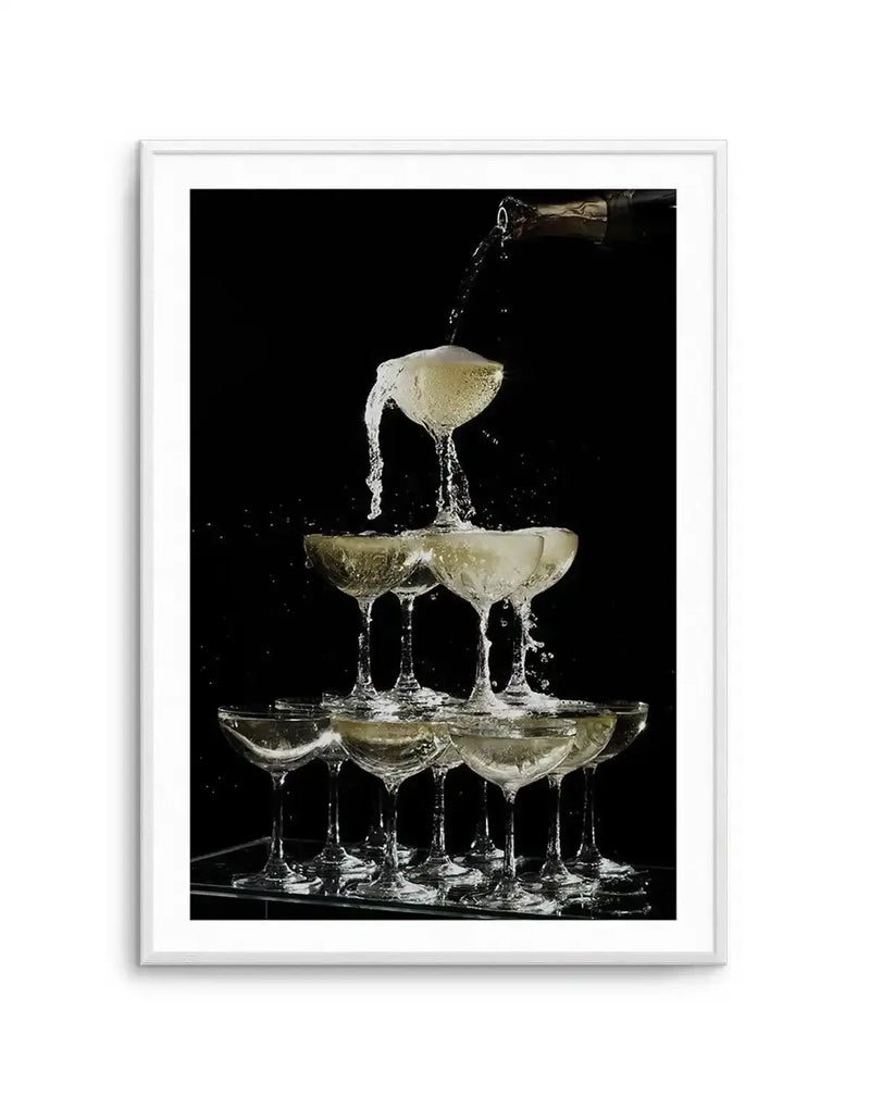 Framed photography art print capturing a champagne fountain in action against a stark black background, with a white frame and white border. A stream of champagne pours from a bottle into the top glass, overflowing and cascading down a pyramid of coupe glasses, creating splashes and frothy bubbles. The golden liquid reflects light, highlighting the elegant arrangement of glasses on a reflective surface.