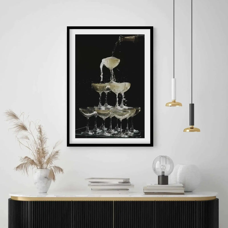 Lifestyle shot of a framed art print displayed above a black console table in a modern room. The print features a champagne fountain with sparkling liquid pouring from a bottle into a top glass, cascading down into multiple coupe glasses arranged in a pyramid, set against a dark background. The room includes a white marble top on the console, dried pampas grass in a white vase, stacked books, a clear globe lamp, a white ribbed vase, and two pendant lights with gold and white/black accents.