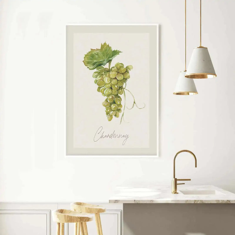 Lifestyle shot of a kitchen with a framed art print displayed on a white wall above a counter. The art print features a watercolour illustration of a bunch of green Chardonnay grapes with a single green leaf, on a cream background, with a white frame and white border. The kitchen includes a white marble countertop, a gold faucet, two hanging pendant lights with white shades and gold interiors, and two light wood bar stools.