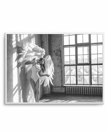 Framed black and white photograph of a woman with large white angel wings standing in a sunlit room. The woman, wearing a light-colored bodysuit, stands against a textured wall with strong shadows, looking towards a large multi-paned window that reveals a city skyline.