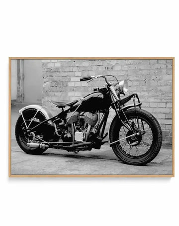 Black and white framed canvas art print featuring a vintage motorcycle photographed from the side against a white brick wall background. The motorcycle has a classic cruiser design with a sidecar attachment, detailed engine components, spoke wheels, and an illuminated headlight. The image captures fine mechanical details and textures of the bike. The canvas is mounted in a natural wood frame with a light tan finish.