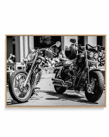 Black and white framed canvas art print depicting three classic motorcycles parked side by side on a street. The motorcycles feature chrome details, whitewall tires, and vintage styling with prominent headlights and handlebars. The photograph has high contrast monochromatic tones with sharp detail on the bikes and blurred urban architecture in the background. The canvas is mounted in a natural wood frame with a white border, creating a gallery-style presentation.