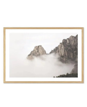 Framed photography art print featuring a misty mountain landscape with a natural oak frame and white border. The composition shows rugged, rocky mountains partially obscured by thick, soft white clouds or fog, creating an ethereal and serene mood.