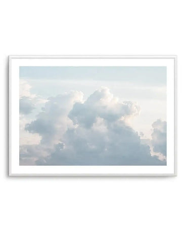 Framed photography art print featuring a sky filled with fluffy white and pale blue cumulus clouds, with soft sunlight illuminating their edges, displayed in a white frame with white border.