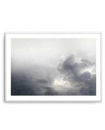 Framed black and white photograph of a dramatic cloudy sky with a white frame and white border. The composition features dark, turbulent storm clouds on the right, with bright light breaking through in the center, contrasting with lighter, hazy clouds on the left.