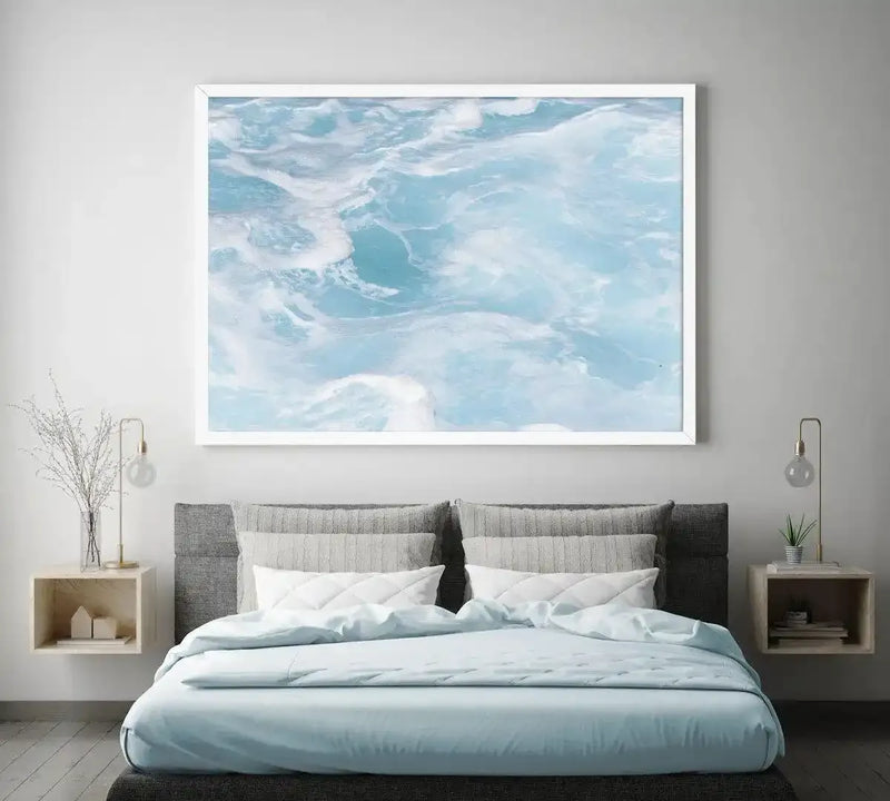 Framed art print depicting an aerial view of turquoise ocean waves with white foam and seafoam patterns. The photograph shows swirling water formations in shades of pale blue, aqua, and white against a light gray background. The print is mounted in a white frame with white border and displayed above a modern bed with gray upholstered headboard, light blue bedding, and two wooden nightstands with pendant lights in a minimalist bedroom setting.