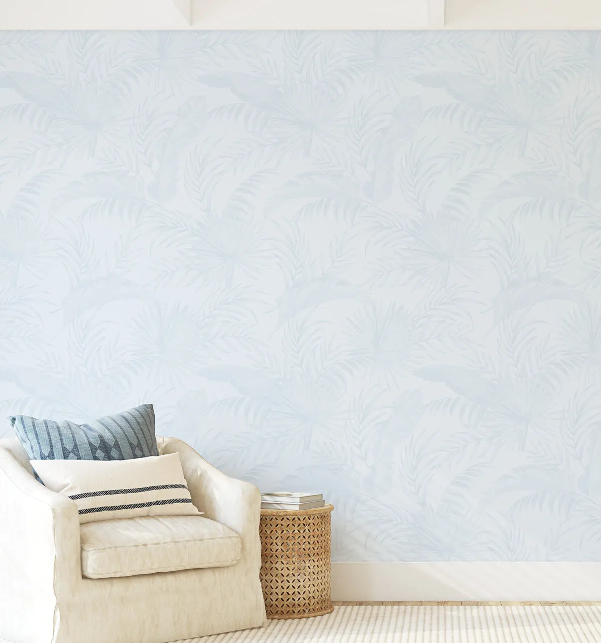 Peel and stick wallpaper featuring a repeating pattern of hand-painted tropical palm leaves in a soft duck egg blue on a light blue background. The botanical design has a loose, organic watercolour quality with visible brushstroke texture. A cream armchair with blue and cream striped cushions and a woven side table with books are in the foreground.
