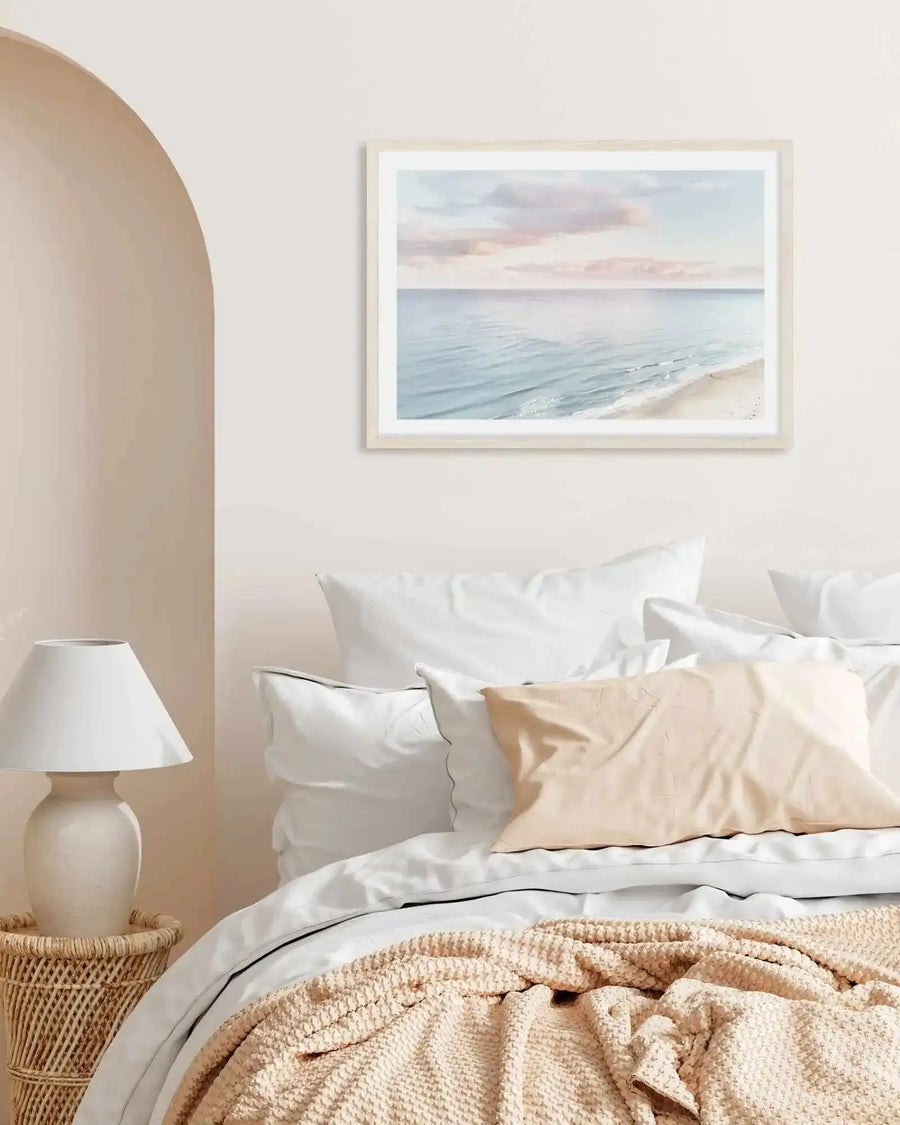 Lifestyle shot of a bedroom with a framed art print of a coastal sunset above a bed. The print features a serene beach scene with gentle blue-green waves lapping at a sandy shore under a sky with soft pink and pale blue clouds, displayed in a natural wood frame with a white border. The bed is dressed with white linens, a light peach textured throw, and a peach pillow. To the left, a white lamp sits on a woven rattan side table, next to a wall with a soft peach arched alcove.