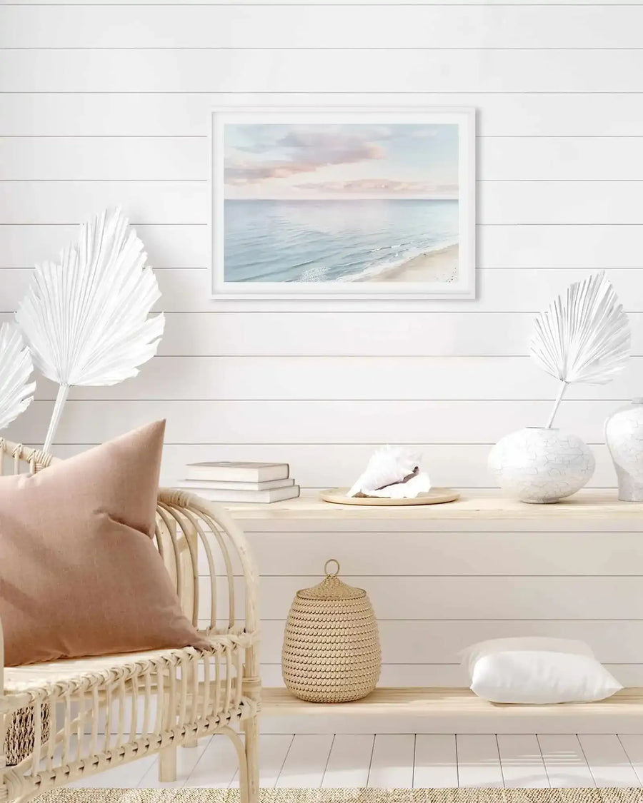 Pastel coastal seascape in white frame on white shiplap wall above rattan chair in coastal room