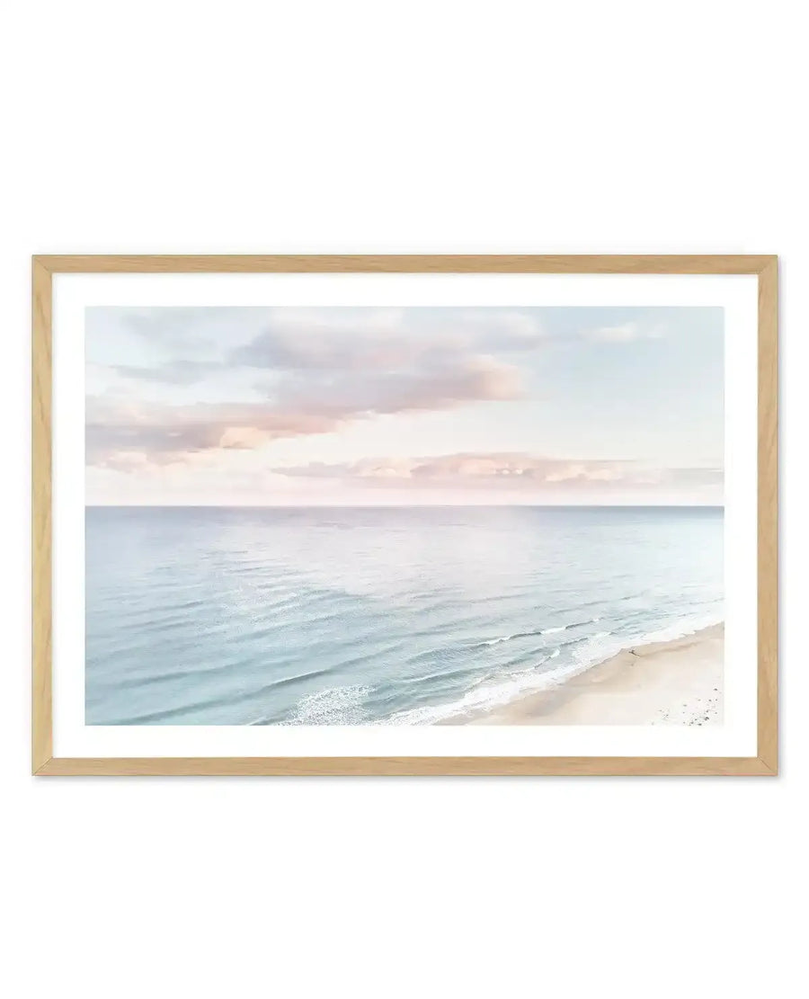Framed photography art print of a serene coastal beach scene at sunset, with a natural wood frame and white border. The composition features gentle, light blue waves breaking on a sandy beach, with a vast expanse of calm ocean stretching to the horizon under a soft sky with pastel pink and light blue clouds.