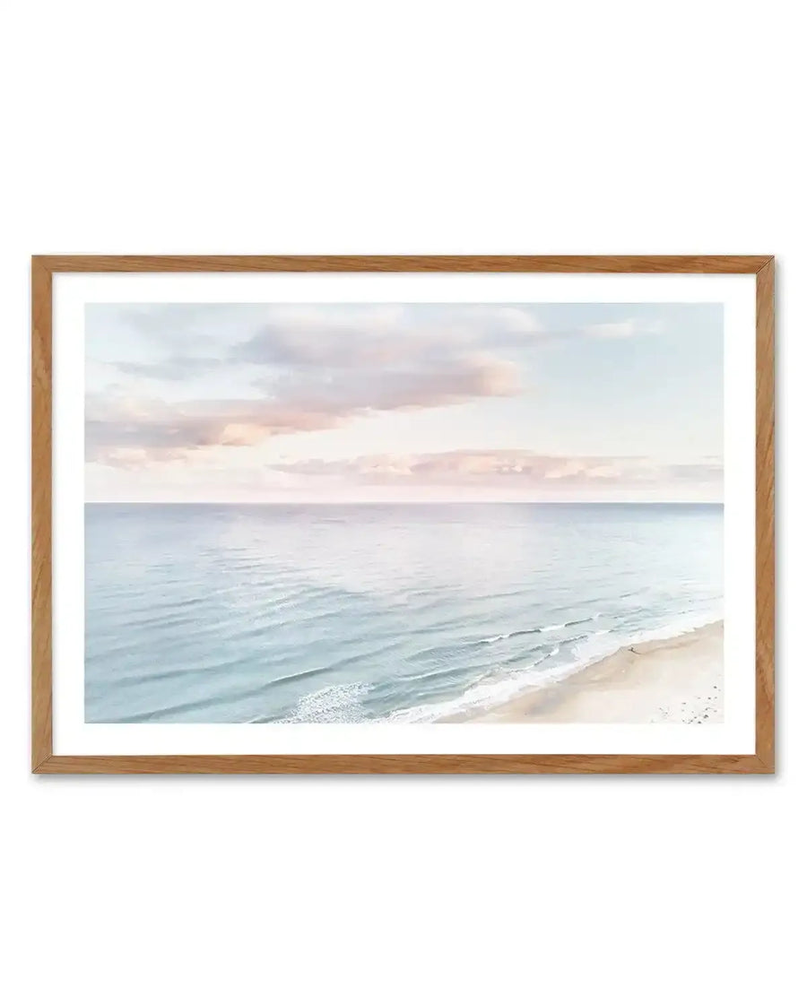 Framed photography art print of a coastal beach scene at sunset with natural wood frame and white border. The composition features a wide expanse of calm ocean with gentle waves breaking on a sandy beach, under a sky with soft pink and blue clouds.