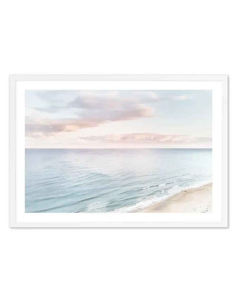 Framed photography art print of a coastal beach scene with a white frame and white border. The composition features a calm ocean with gentle waves meeting a sandy beach under a pastel sky with soft pink and blue clouds.