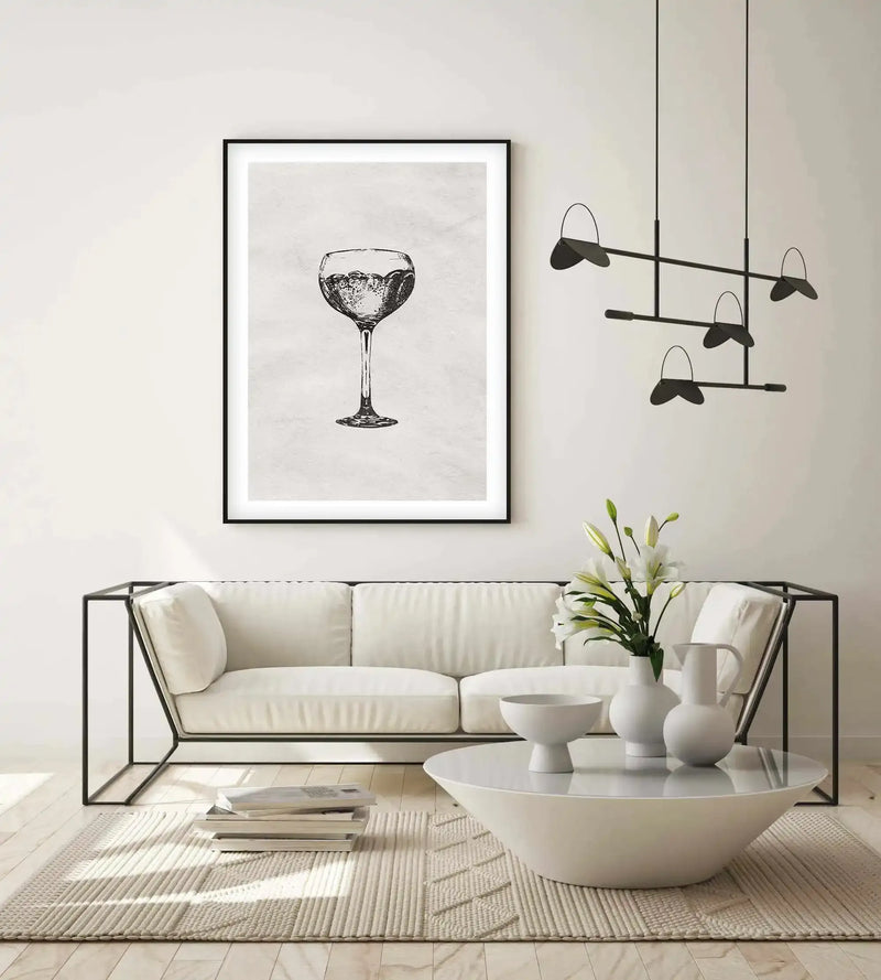 Black-framed art print displayed above a cream upholstered sofa in a minimalist living room. The print features a detailed pen-and-ink illustration of a coupe cocktail glass filled with sparkling liquid, centered on a white background with subtle gray texture. The artwork is matted with a white border within the black frame. Below, a round white pedestal table holds white ceramic vessels and fresh greenery, accompanied by a neutral striped rug and modern black mobile sculpture hanging above.
