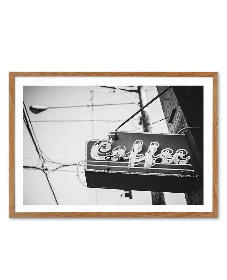 Framed black and white photograph of a vintage "Coffee" neon sign attached to a brick building, with a natural wood frame and white border. The sign features the word "Coffee" in a cursive script, with visible neon tubing. Utility poles and wires are visible in the background against a bright sky.