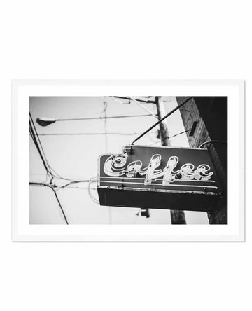 Framed black and white photograph of a vintage "Coffee" neon sign attached to a brick building, with a natural wood frame and white border. The sign features stylized, cursive lettering that appears worn and slightly rusted, with visible wiring and support bars. In the background, power lines and a streetlamp are visible against a bright, overcast sky, creating a nostalgic urban scene.