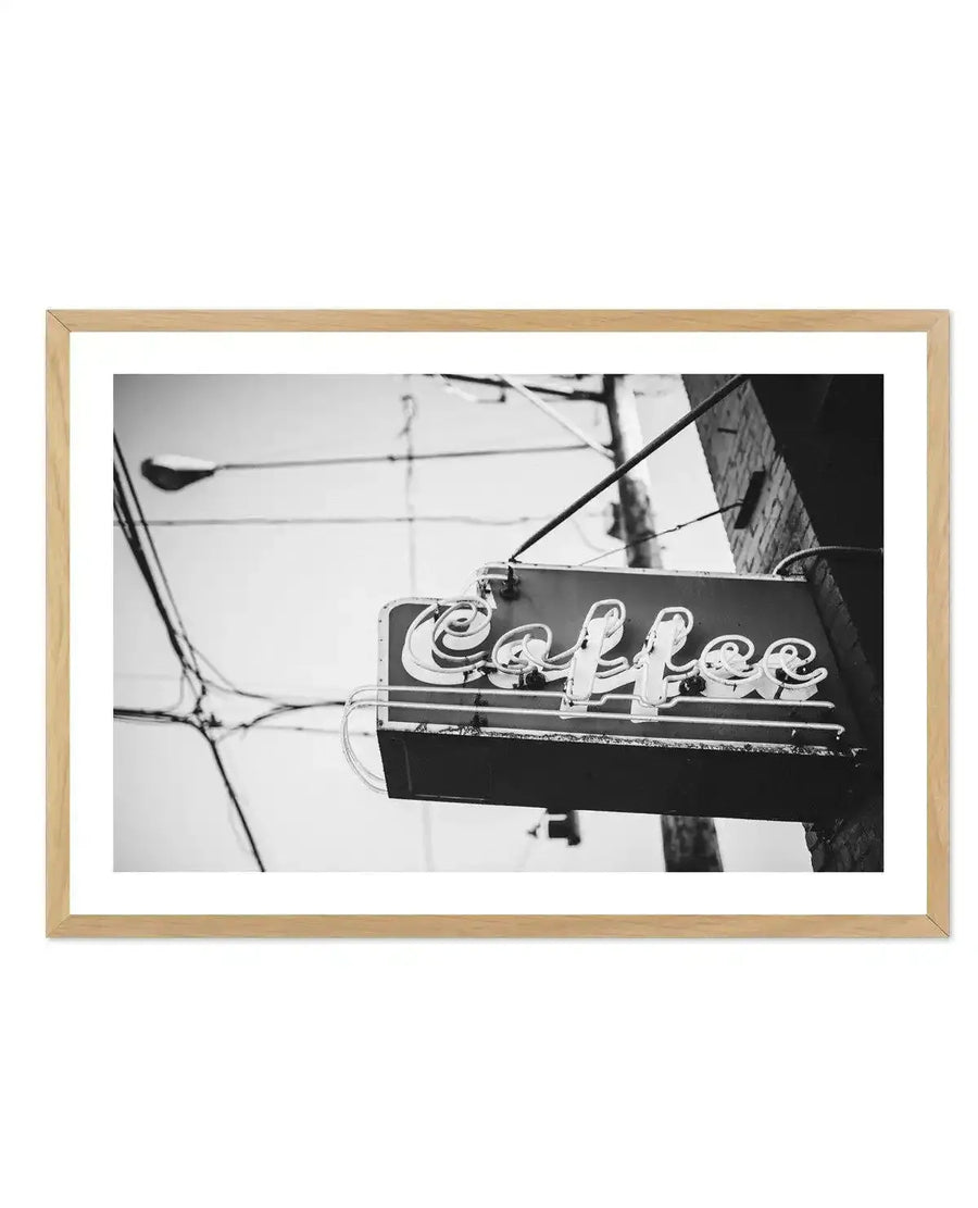 Framed black and white photograph of a vintage "Coffee" neon sign attached to a brick building, with a natural wood frame and white border. The sign features stylized, cursive lettering, with some of the neon tubes appearing broken or unlit, showing wear and age. Utility poles, wires, and a street lamp are visible in the background against a bright, overcast sky.