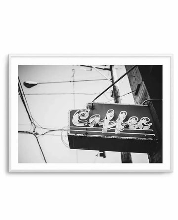 Framed black and white photograph of a vintage "Coffee" neon sign hanging outdoors. The sign features stylized, glowing white letters against a dark rectangular background, suspended by wires and brackets from a brick building. Utility poles and a street lamp are visible in the soft focus background, creating a nostalgic urban scene.
