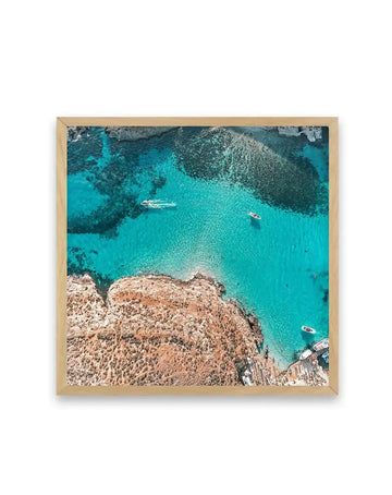 Aerial photograph printed on art paper in natural wood frame showing turquoise Mediterranean waters surrounding rocky tan and grey outcroppings, with several small white and red boats anchored in the shallow cove. The composition captures the contrast between the textured limestone cliffs in the foreground and the crystalline blue-green water dotted with darker seagrass beds. The natural wood frame complements the coastal landscape imagery.
