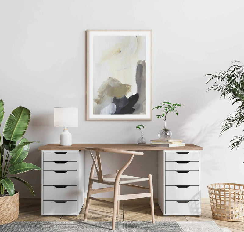 Lifestyle shot of a home office with a framed abstract art print displayed above a wooden desk. The print features large, expressive brushstrokes in warm cream, muted beige, and deep charcoal grey, with a natural wood frame and white border. The desk has white drawer units on either side, a white lamp, and small potted plants. A light wood chair with a woven seat is in front of the desk, and large green plants are on either side of the room.