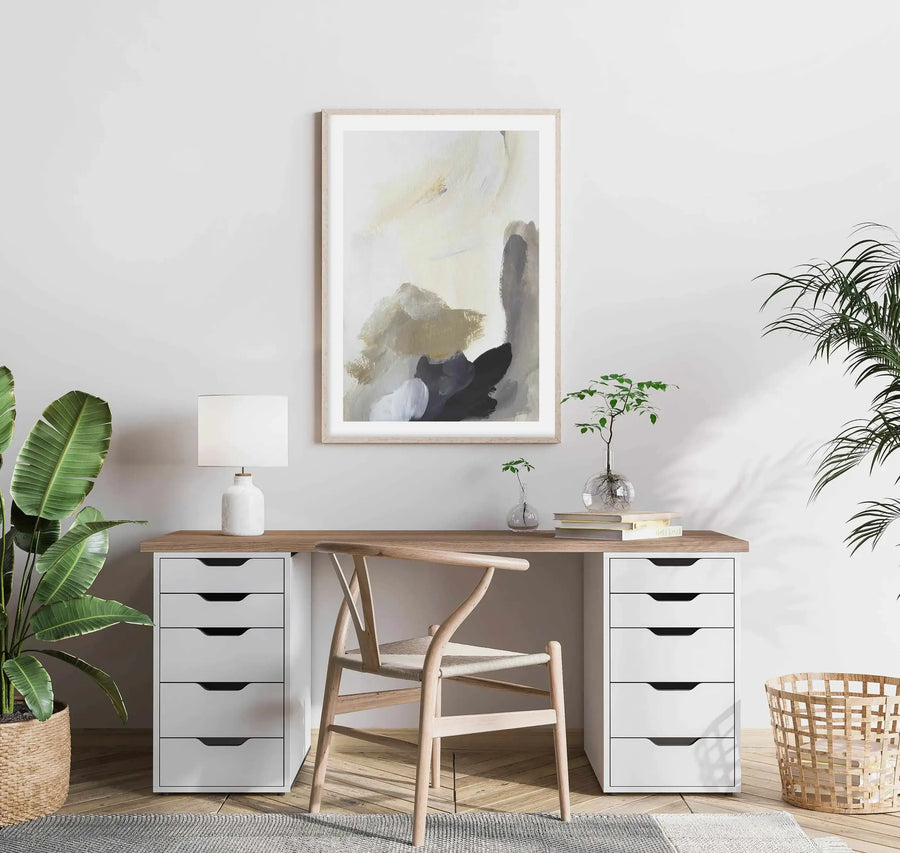 Lifestyle shot of a home office with a framed abstract art print displayed above a wooden desk. The print features large, expressive brushstrokes in warm cream, muted beige, and deep charcoal grey, with a natural wood frame and white border. The desk has white drawer units on either side, a white lamp, and small potted plants. A light wood chair with a woven seat is in front of the desk, and large green plants are on either side of the room.