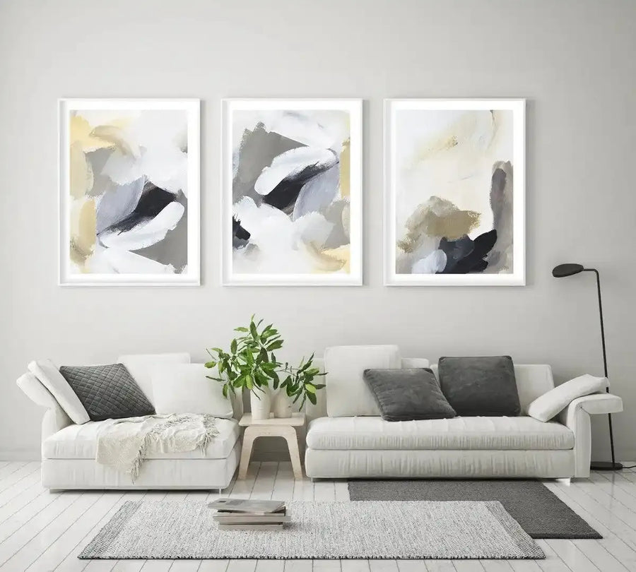 Three framed abstract art prints displayed above a white sectional sofa in a minimalist living room. The prints feature swirling brushstrokes in shades of white, light grey, charcoal, and pale yellow, creating dynamic compositions. The room includes a light grey textured rug, a small wooden side table with a plant, and dark grey throw pillows.