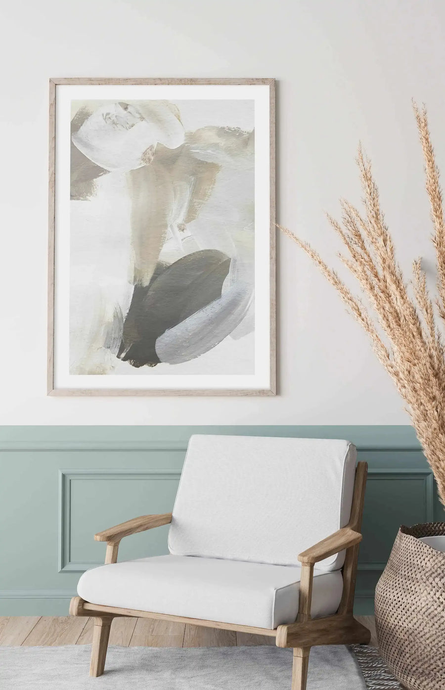Lifestyle shot of a contemporary art print displayed above a white armchair in a living room with a two-tone wall. The art print features abstract brushstrokes in white, beige, grey, and black, with a natural wood frame and white border. The lower half of the wall is painted a muted teal, and a woven basket with dried pampas grass is visible on the right.