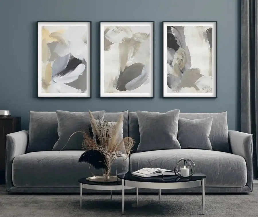 Lifestyle shot of a living room featuring three framed abstract art prints displayed above a grey velvet sofa. The prints feature swirling brushstrokes in varying shades of grey, white, cream, and pale yellow, creating dynamic compositions. The room includes a dark wood side table, a coffee table with a candle and book, and a vase of dried pampas grass.