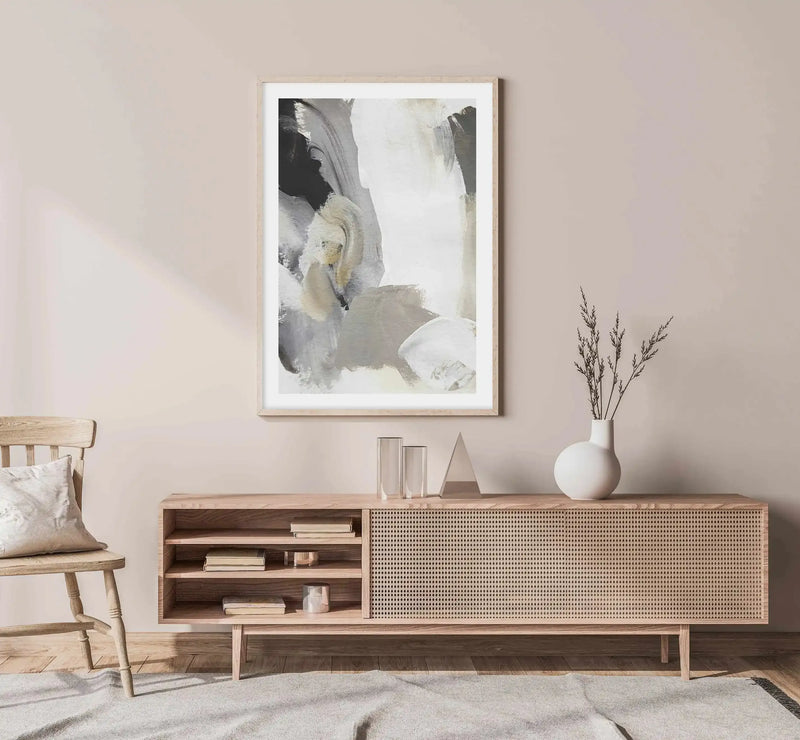 Large framed abstract art print displayed above a light wood console table in a minimalist living room. The print features broad, expressive brushstrokes in shades of black, white, grey, and beige, creating a dynamic, layered composition. The console table has open shelving with books and decorative objects, and a woven cane front. A light wood chair with a cream cushion is to the left, and a white vase with dried grasses sits on the table to the right. A light grey rug covers the floor.