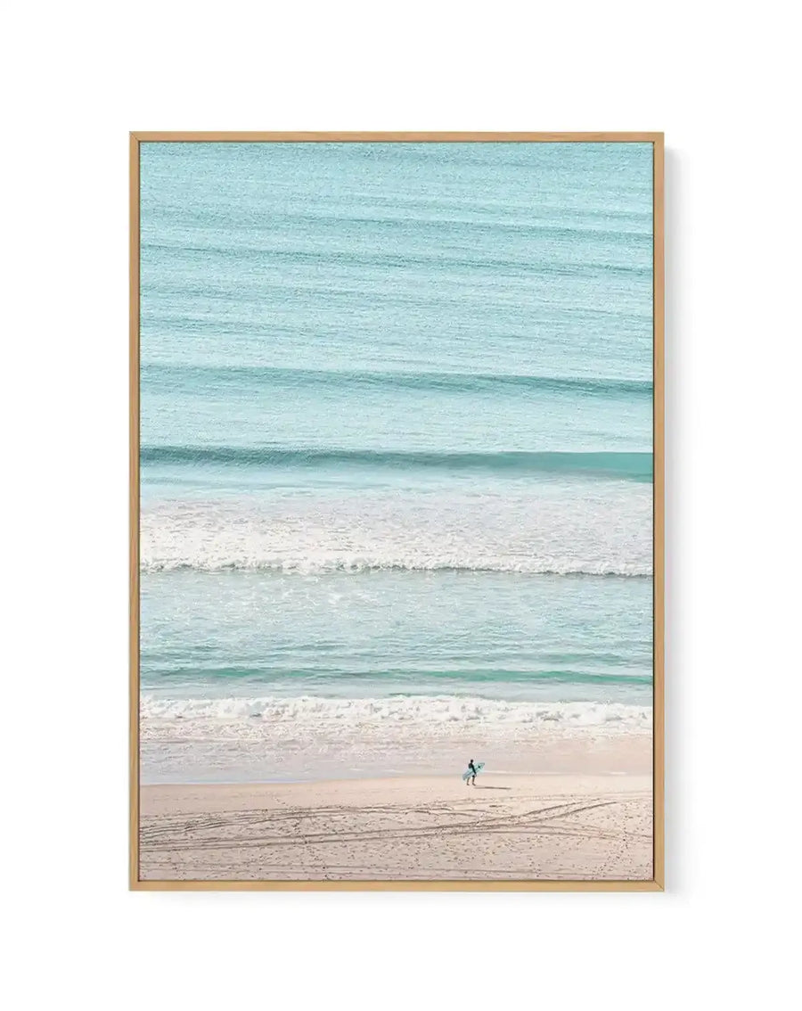 Framed canvas art print featuring a serene beach scene with horizontal bands of colour: pale turquoise sky at top, deeper turquoise ocean with gentle waves in the middle, pale sand beach, and a solitary surfer in a wetsuit standing on the shore holding a surfboard. Natural wood frame with white border. Soft, photographic style with muted, coastal tones creating a calm, minimalist composition.