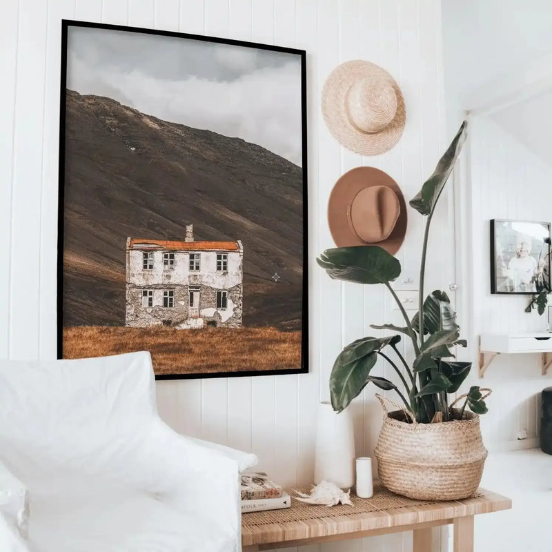 Framed art print displayed on a white shiplap wall showing a cream and rust-colored cottage with orange roof nestled in a vast, dark brown mountainous landscape under an overcast sky. The photograph has warm, muted tones with golden-brown grass in the foreground. Black frame with white border. Styled with a woven basket containing green potted plant, cream straw hats, and white ceramic items on a light wood shelf below.