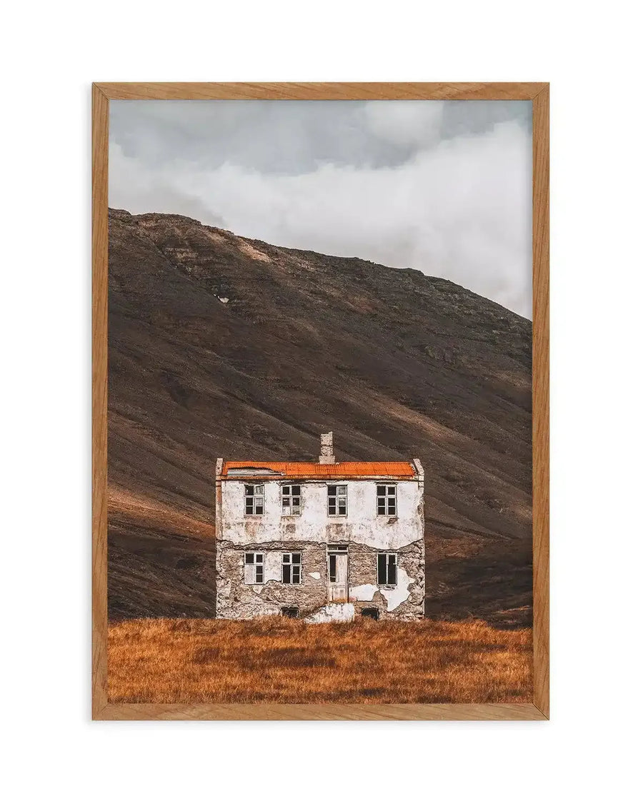 Framed art print featuring a photograph of an abandoned two-story stone house with a bright orange roof, set against a barren dark brown mountainside. The house has weathered white-painted upper walls, stone lower walls, and multiple windows. Golden-brown grass covers the foreground. Overcast gray and white sky fills the top. Natural wood frame with visible grain texture surrounds the image.