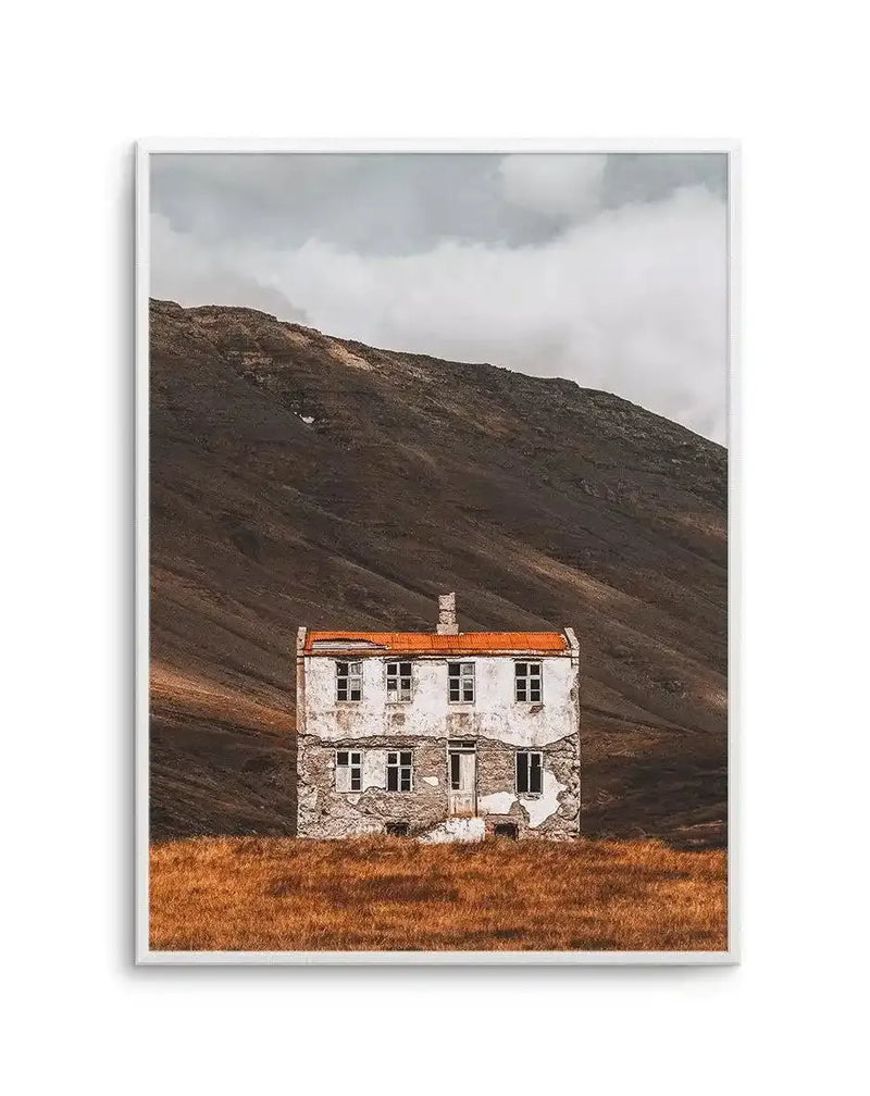 Framed art print featuring a photograph of an abandoned two-story stone house with white-painted upper walls and orange-tiled roof, standing alone in a barren landscape. The foreground shows golden-brown dried grass, while a dark, rocky mountain slope dominates the background under an overcast sky with white and gray clouds. The image has a muted, cool color palette with earthy browns and whites. Presented in a silver-toned frame with white border.