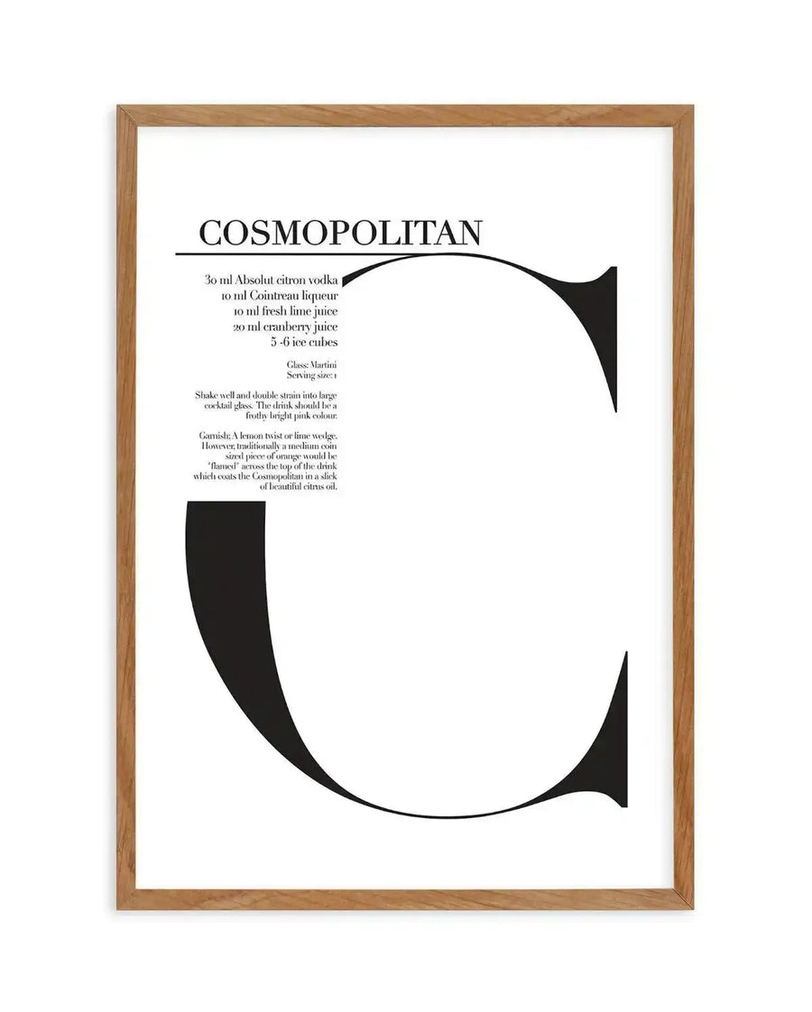 Art print featuring the word "COSMOPOLITAN" in a bold, elegant serif font at the top, with a large, stylized black capital "C" dominating the lower two-thirds of the white background. The recipe for a Cosmopolitan cocktail is printed in a smaller, delicate serif font to the left of the "C", detailing ingredients, serving instructions, and garnish. The print is displayed in a natural oak frame with a white border.