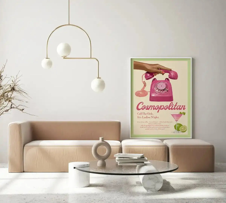 Large framed art print displayed in a minimalist living room above a low-profile sofa. The print features a vintage-style illustration of a hand holding a bright pink rotary telephone, with the text "Cosmopolitan Call The Girls, It's Ladies Night" and a pink cocktail with lime wedges. The print has a white frame and a pale green border. The room includes a beige sectional sofa, a glass coffee table, and a modern gold chandelier.
