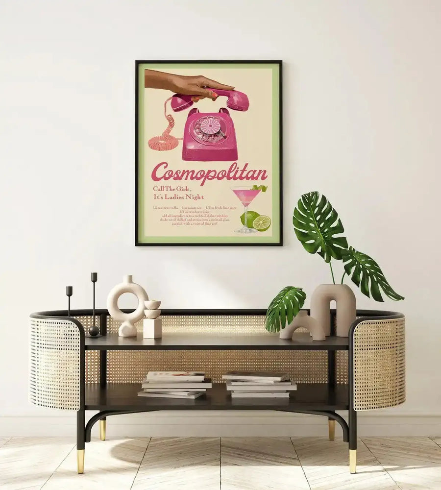 Framed art print displayed above a dark wood and rattan console table in a minimalist living room. The print features a retro-style illustration of a hand holding a pink rotary telephone, with a Cosmopolitan cocktail and lime wedges below, against a pale green background, with a black frame and white border. The room includes white ceramic decorative objects and a large green monstera plant in a beige vase.