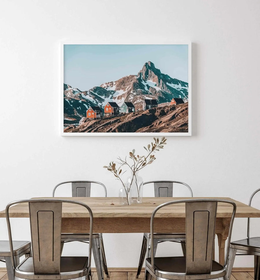 Framed art print displayed on a white wall above a wooden dining table with metal chairs. The print shows a landscape photograph featuring colorful cottages in shades of red, orange, and cream clustered on a rocky, brown hillside. Behind them rises a dramatic snow-capped mountain peak against a pale blue sky. The photograph has a white frame and white border, creating a clean, minimalist presentation in a contemporary dining space with a clear glass vase of dried botanical stems on the table.