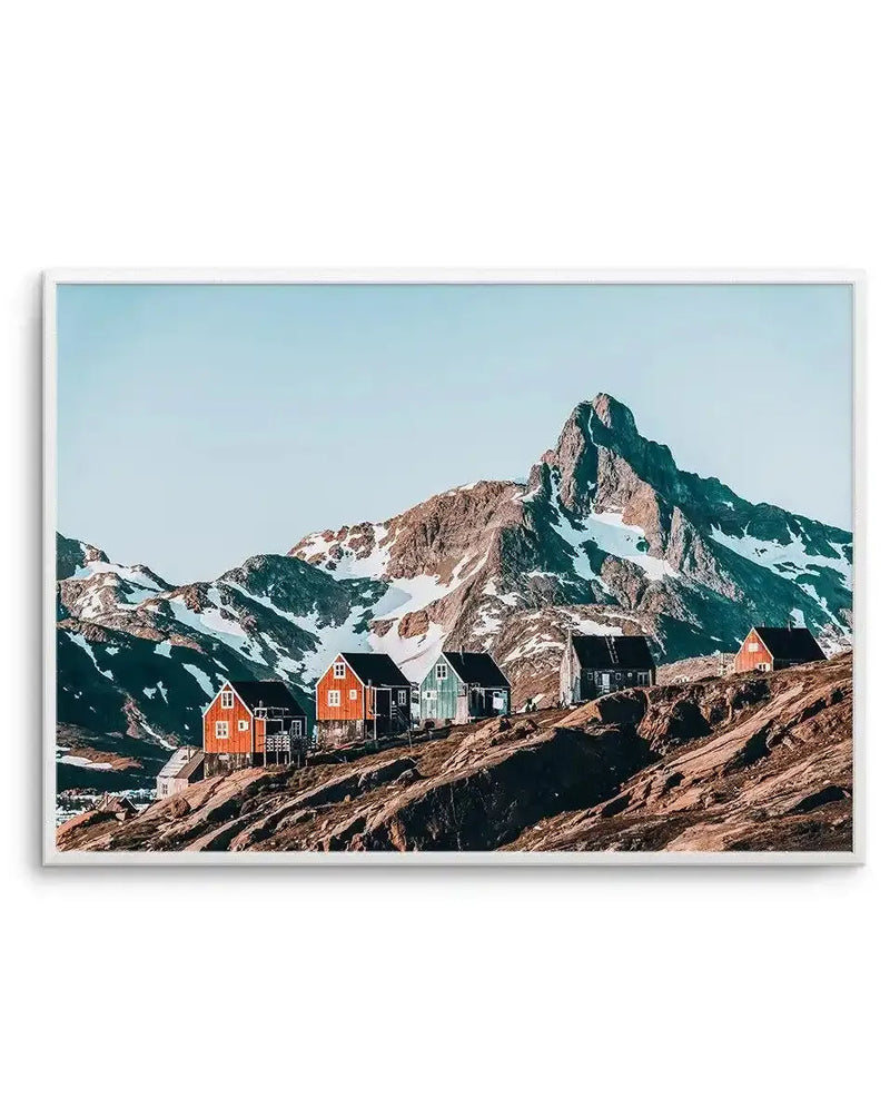 Framed art print featuring a photograph of colorful cottages nestled on rocky terrain beneath a dramatic snow-capped mountain peak. The cottages are painted in shades of red, orange, green, and black with dark roofs. The mountain dominates the background with exposed brown and tan rock faces and patches of white snow. Clear blue sky fills the upper portion. The photograph has rich, saturated colors and crisp detail. The print is displayed in a white frame with a white border.
