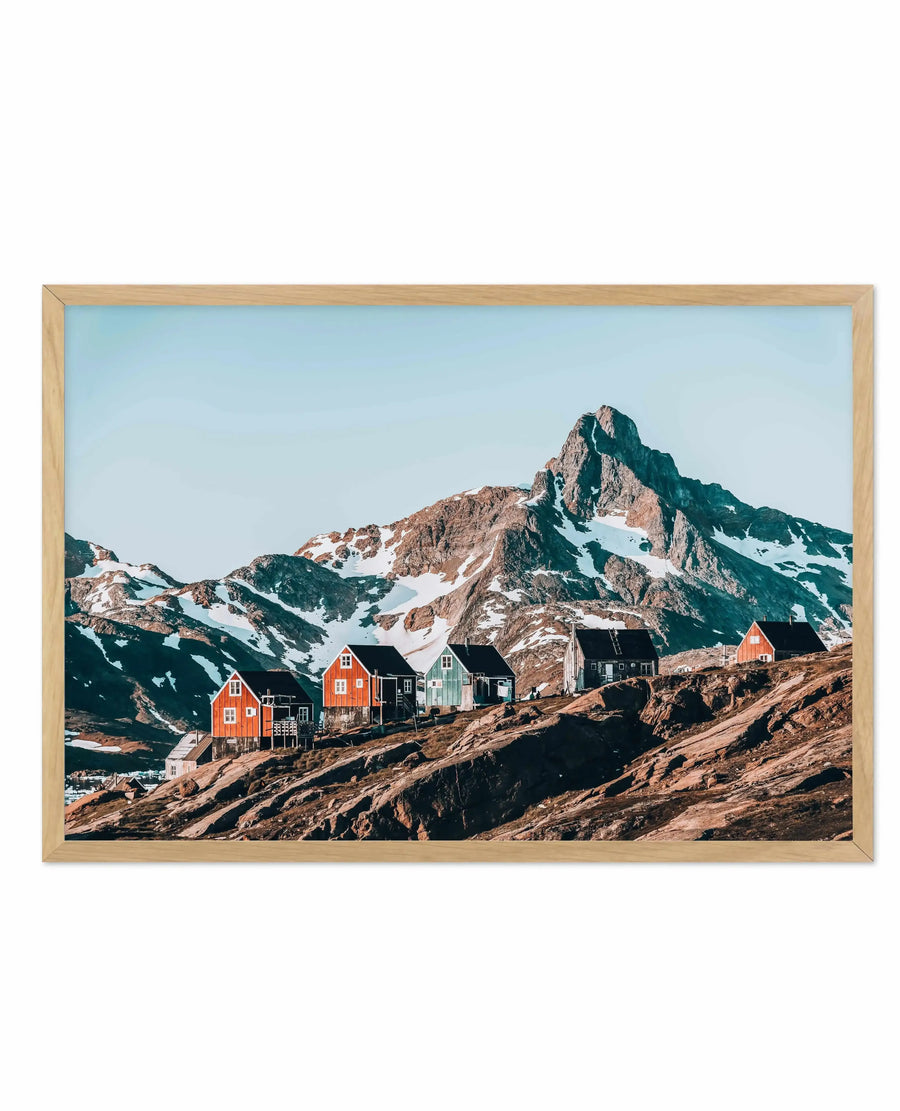 Framed art print featuring a photograph of colorful houses nestled on rocky terrain beneath a snow-capped mountain peak. The composition shows six residential buildings in bright red, black, white, and green situated on brown rocky slopes. A majestic mountain with snow patches and green-tinted upper slopes dominates the background under a clear light blue sky. The natural wood frame has a warm tan finish and displays the landscape photograph with a white border.