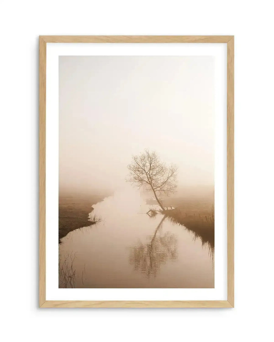 Framed art print featuring a misty landscape photograph with a solitary bare tree leaning over still water. The composition shows muted tones of beige, taupe, and soft cream creating a foggy atmosphere. The leafless tree is reflected in calm water below, with marshland visible along the banks. Natural wood frame with white border surrounds the print, creating a serene, minimalist aesthetic with soft, diffused lighting throughout.