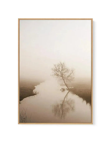 Framed canvas art print featuring a serene misty landscape with a bare tree standing on the bank of a still water body. The composition shows a calm river or stream reflecting the pale sky and tree, with soft beige, cream, and taupe tones throughout. Sparse grasses line the foreground while fog obscures the distant banks, creating a tranquil, minimalist atmosphere. The artwork has a soft, photographic quality with muted earth tones. Natural wood frame with subtle grain texture.