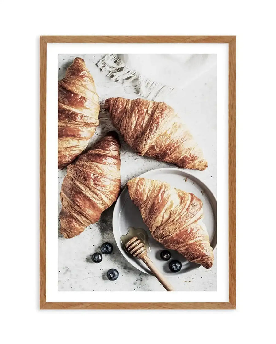 "Framed photography art print featuring four golden-brown croissants on a light grey textured surface, displayed in a natural oak frame with white border. One croissant rests on a small white plate with a honey dipper and a drizzle of honey, surrounded by scattered blueberries."