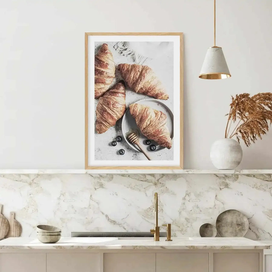 Lifestyle shot of a kitchen featuring a framed art print of croissants displayed above a marble countertop. The print features four golden-brown croissants with flaky texture, one on a white plate with blueberries and a honey dipper, against a light grey background. The room includes a gold faucet, a white ceramic vase with dried pampas grass, and a modern pendant light with a white and gold shade.