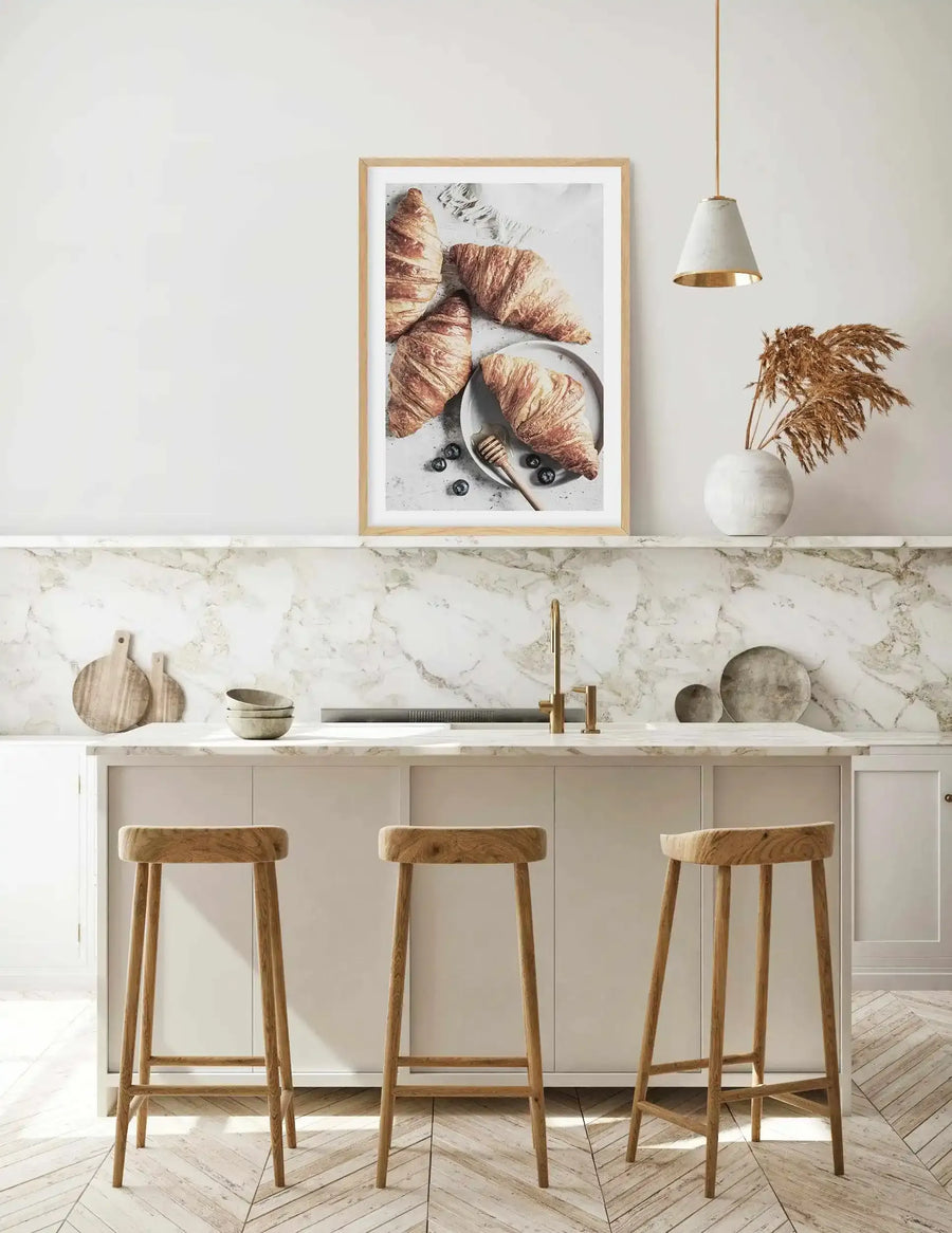Lifestyle shot of a modern kitchen with a framed art print of croissants displayed above a marble backsplash. The print features four golden-brown croissants on a light grey surface, with one croissant on a small white plate next to a honey dipper and scattered blueberries. The kitchen has white cabinets, a gold faucet, and three wooden bar stools. A white vase with dried pampas grass and a white pendant light with a gold interior are also visible.