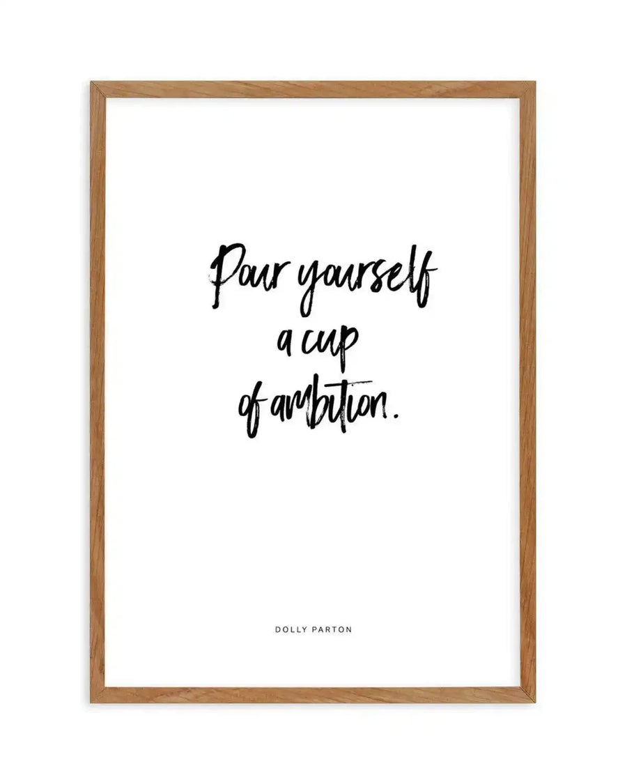 Art print featuring the handwritten quote "Pour yourself a cup of ambition." in black script on a white background, with "DOLLY PARTON" in smaller black capital letters below. The print is displayed in a natural oak frame with a white border.