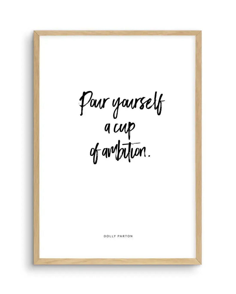 Art print featuring the quote "Pour yourself a cup of ambition." in black handwritten script on a white background, printed on fine art paper and displayed in a natural oak frame with white border. The quote is attributed to Dolly Parton in smaller black text at the bottom.