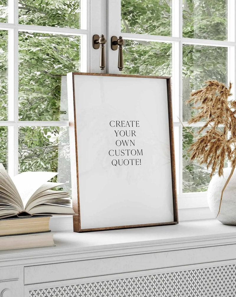 Custom quote art print displayed on a white windowsill in a wooden frame with a white border. The print features centered black serif text reading "CREATE YOUR OWN CUSTOM QUOTE!" on a white background. Styled alongside stacked books, a potted white ceramic vase with dried pampas grass, and a large multi-pane window showing green foliage outside. Natural daylight illuminates the scene.