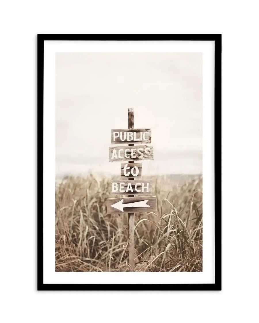 Framed sepia-toned photograph of a weathered wooden signpost in a field of tall, dry grass, with a black frame and white border. The sign has four planks, reading "PUBLIC ACCESS to BEACH" in white block letters, and a fifth plank with a white arrow pointing left. The background shows a hazy horizon line, suggesting a distant body of water under an overcast sky.